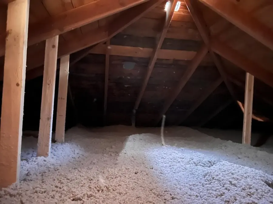 Attic with light-colored blown-in cellulose insulation on the floor under hip roof framing and an old plank gable wall