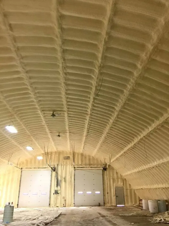 Arched building interior with tan closed-cell spray foam on ceiling and walls, two overhead doors, drums and floor plastic