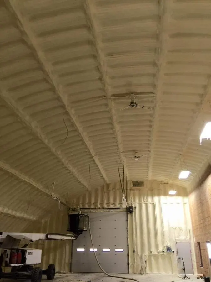 Tan closed-cell spray foam covering the arched ceiling and walls of a metal building with a boom lift and hose
