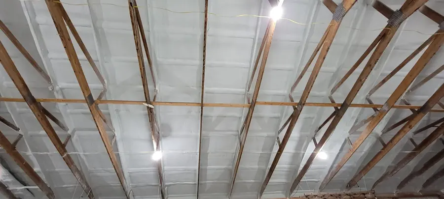 White closed-cell spray foam covering the underside of a roof deck between wood trusses in a large open building