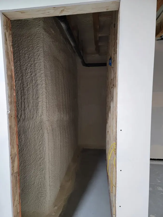 View through a drywalled doorway into a narrow basement closet with a textured tan closed-cell spray foam wall