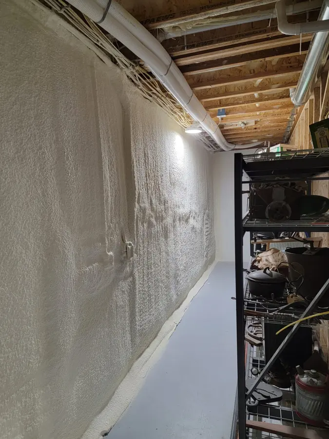 Long basement wall covered in textured white closed-cell spray foam beside a metal storage shelf under exposed floor joists