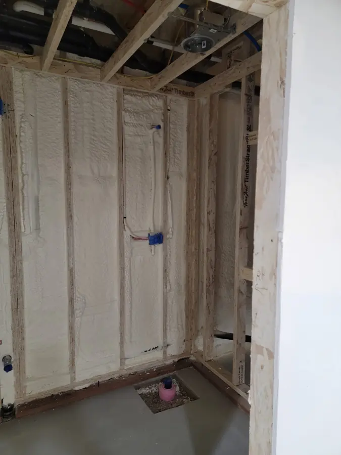 Framed bathroom wall with spray foam in stud bays around plumbing and blue electrical boxes, toilet flange capped on floor
