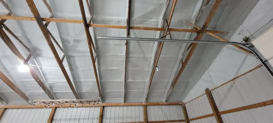 Closed-cell spray foam on the roof deck of a pole barn with an overhead door track and white metal wall panels