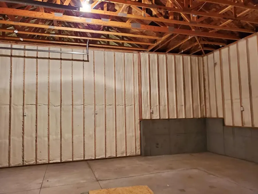 Garage walls with cream closed-cell spray foam filling stud bays above a concrete lower wall, exposed trusses overhead