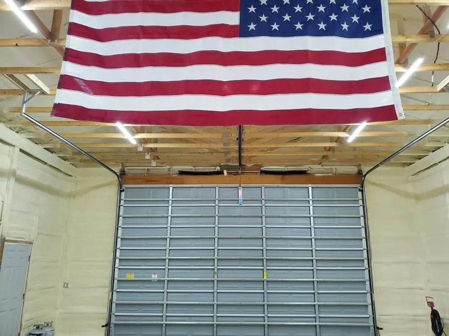Garage with cream closed-cell spray foam on walls, a large American flag above a gray overhead door, and exposed trusses