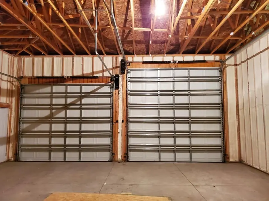 Unfinished garage with spray foam insulation between wall studs and two closed overhead doors under exposed roof trusses