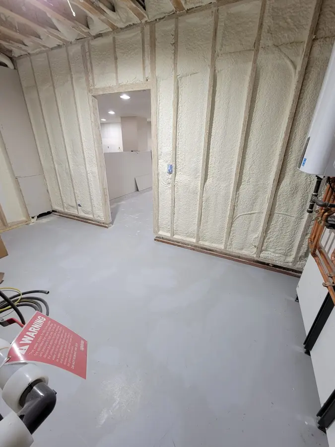 Basement room with spray foam on the walls, gray painted floor, a wall-mounted water heater with copper piping and a warning sign