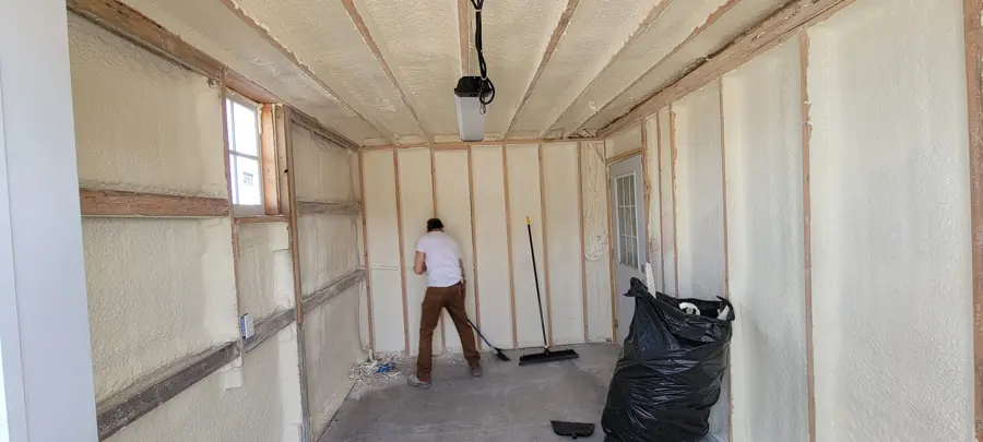 Small building interior with closed-cell spray foam between wall studs and rafters while a worker sweeps the floor