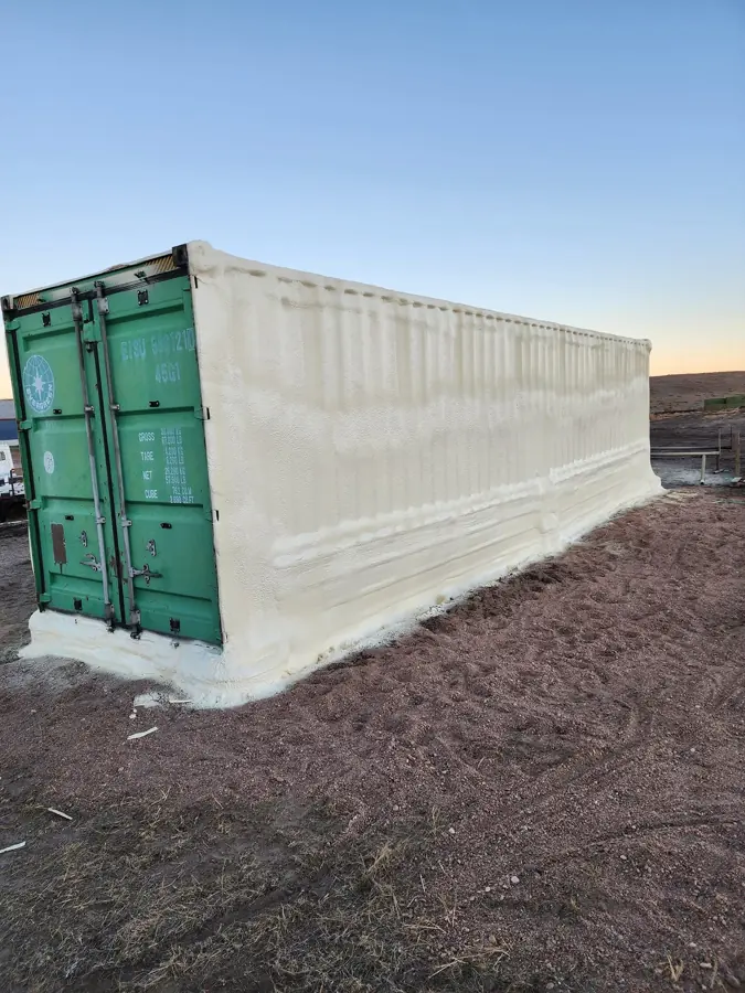 Green shipping container with cream closed-cell spray foam covering the side and roof, on gravel at sunset