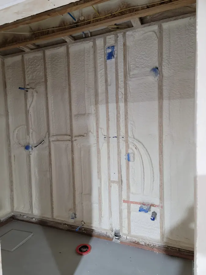 Framed wall with spray foam filling stud bays and covering pipes and wiring, blue electrical boxes and a floor drain cover