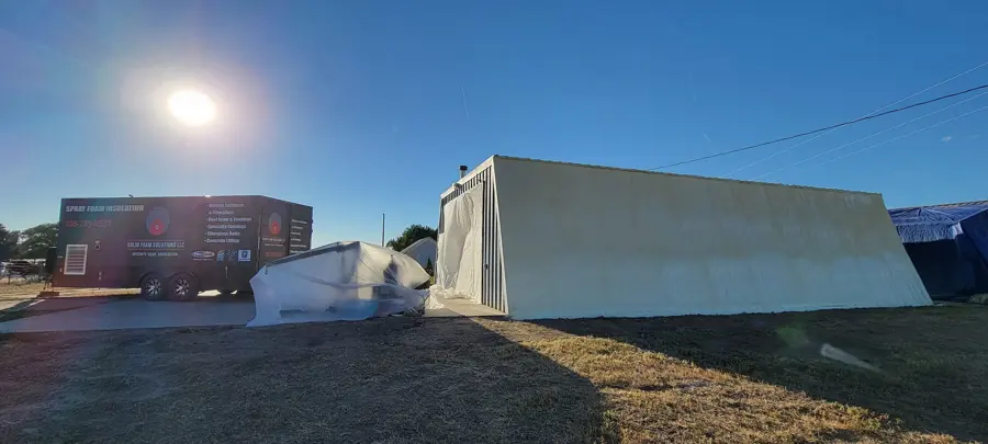 Branded spray foam trailer parked next to a metal building with a cream foam coating, under a low sun