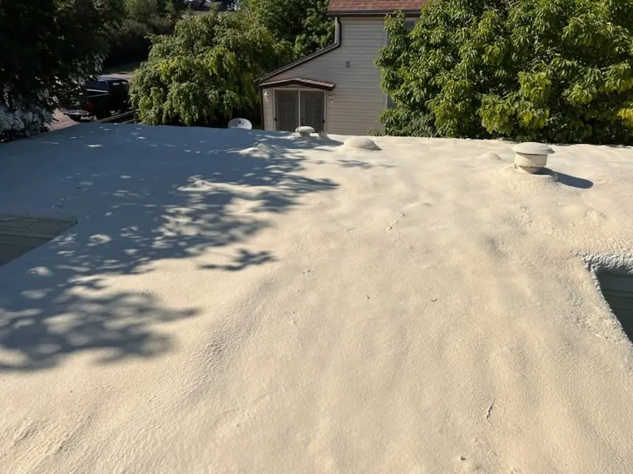 Tan spray polyurethane foam applied over a flat roof with vent caps, with a house and trees behind